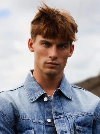 Close-up of a redhead man with a serious expression wearing a denim jacket in an outdoor setting against a cloudy sky backdrop.