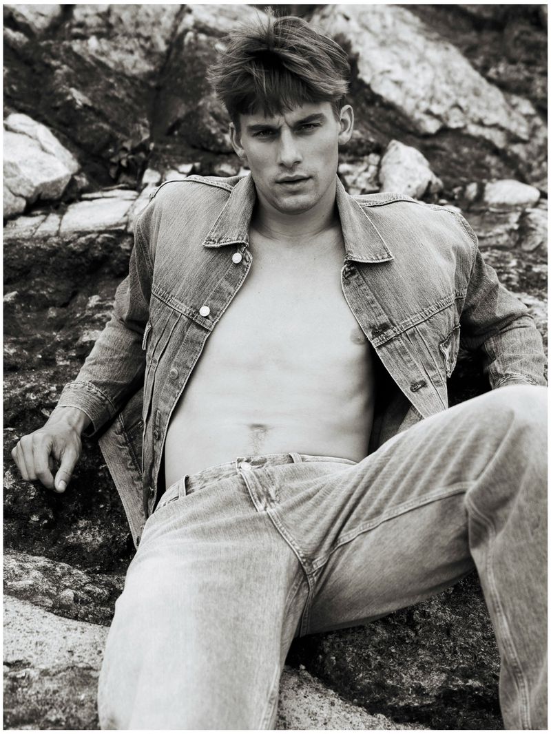 Mikkel Matthiesen in an unbuttoned denim jacket and jeans reclining against coastal rocks in a black and white fashion editorial.