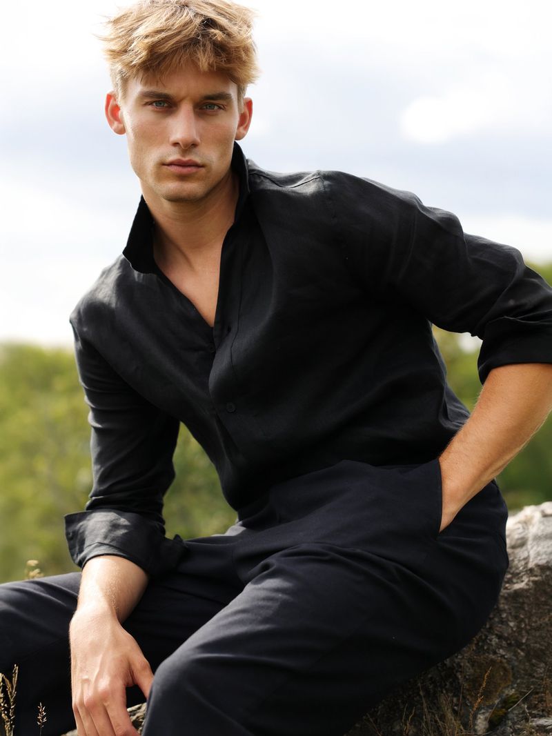 Blonde man in black linen shirt and pants sits outdoors on rock; portrait of male fashion in casual wear on natural backdrop.