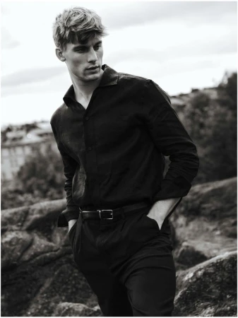 Black and white portrait of a male model in a dark button-down shirt and pants, posing outdoors with a thoughtful expression and hands in pockets.