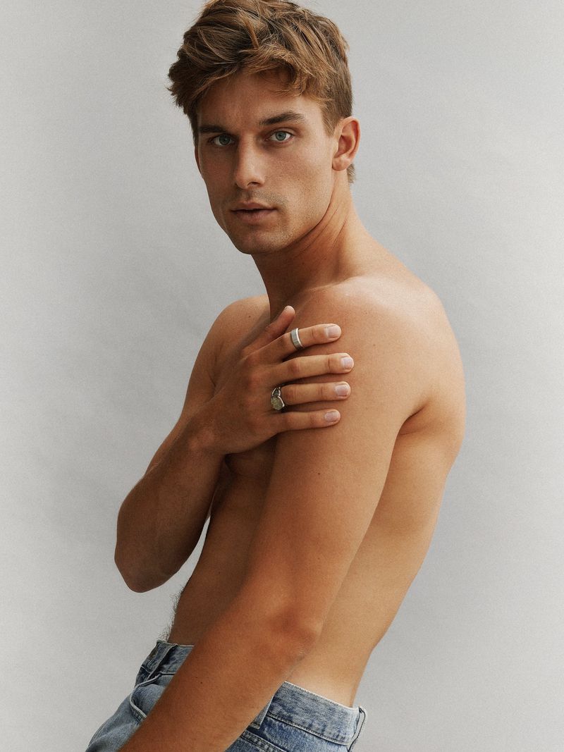 Portrait of a shirtless young man with light brown hair and blue eyes, wearing jeans and rings, against a neutral grey background.