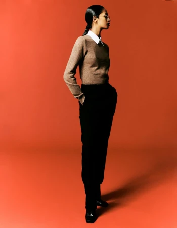 Model Jessie Aina standing in profile wearing a brown crewneck sweater, collared shirt, and black trousers against an orange backdrop.