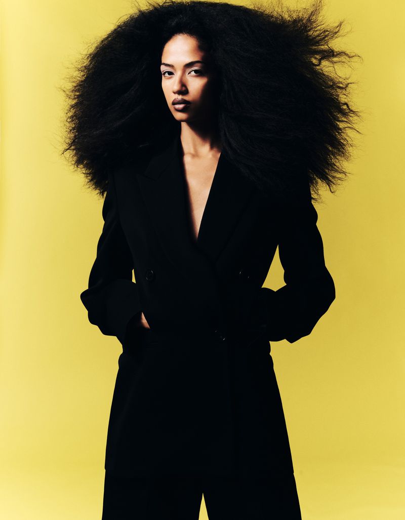 Model with voluminous dark hair wearing a tailored black blazer against a solid bright yellow backdrop.