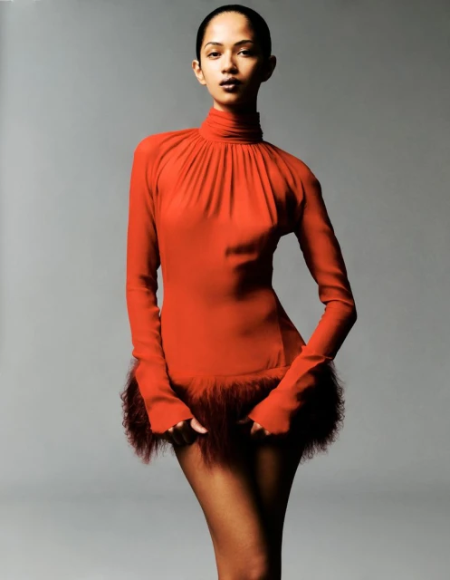 A woman in a red minidress with feathered trim strikes a studio pose, highlighted by a grey backdrop and soft lighting, showcasing fashion and elegance.