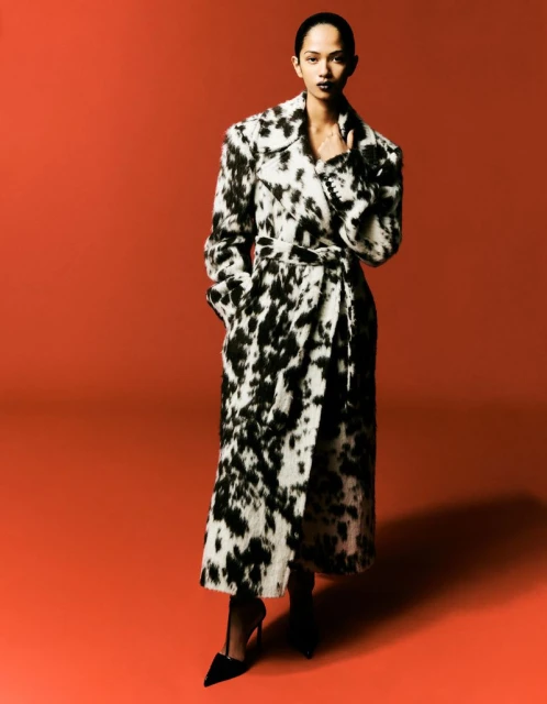 A person with dark hair and dark lipstick, wears a black and white coat with a tie in a photo studio with a red backdrop.