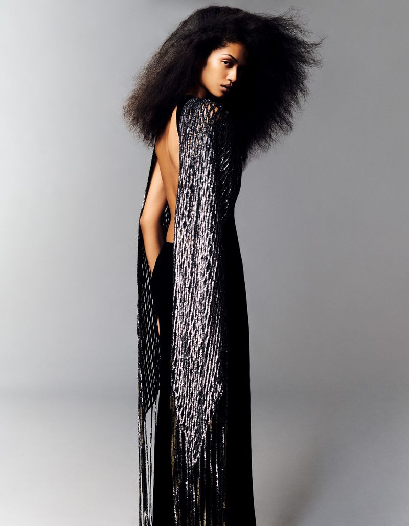 A woman with voluminous dark hair, wearing a black backless gown with long silver fringe, posing against a neutral gray studio backdrop.