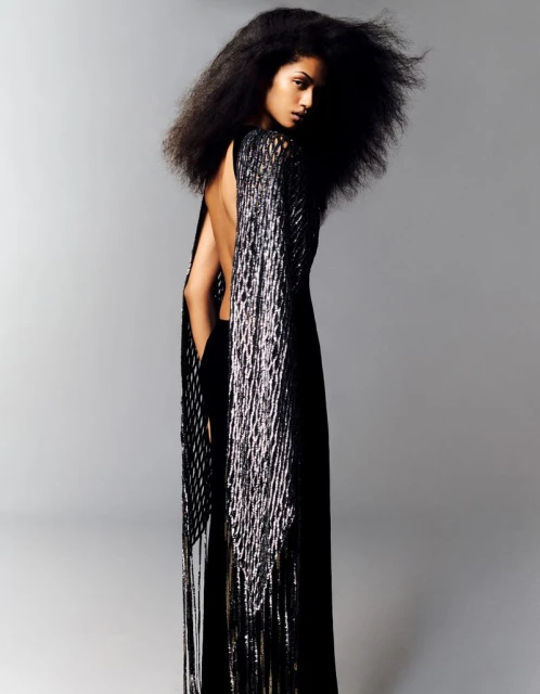 A woman with voluminous dark hair, wearing a black backless gown with long silver fringe, posing against a neutral gray studio backdrop.