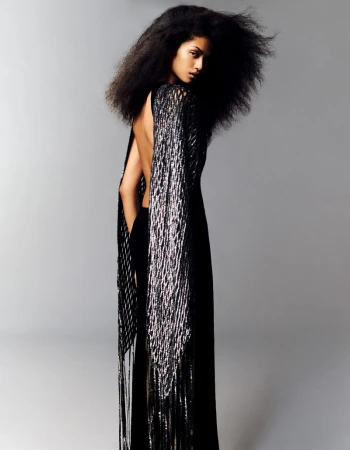 A woman with voluminous dark hair, wearing a black backless gown with long silver fringe, posing against a neutral gray studio backdrop.