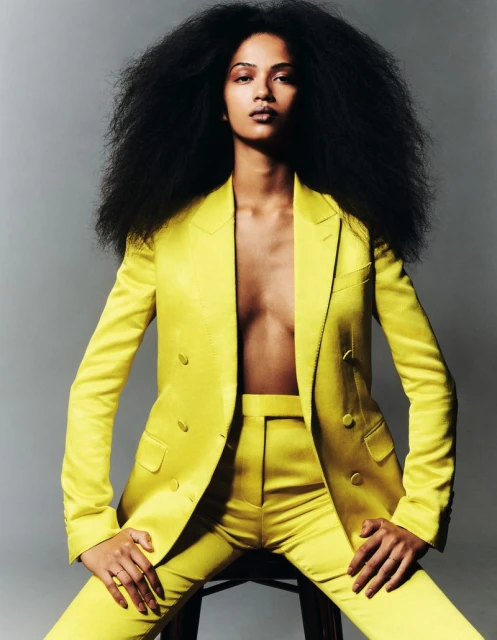 Fashion portrait of woman with afro hairstyle wearing yellow pantsuit. She poses on a stool in front of a grey studio background.