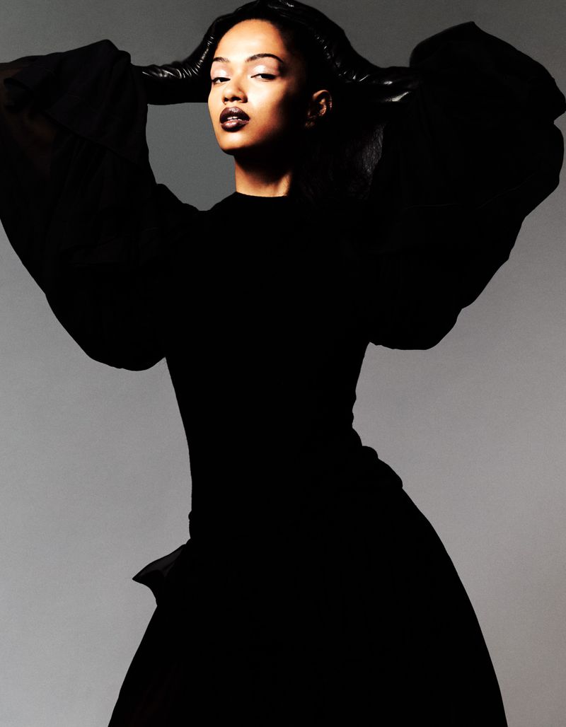 Model wearing a black dress with dramatic puffed sleeves posing against a plain grey studio backdrop