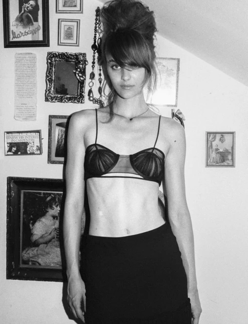 Black-and-white portrait of a young woman with bangs and updo in a bra and skirt, posing indoors against a wall with framed pictures.