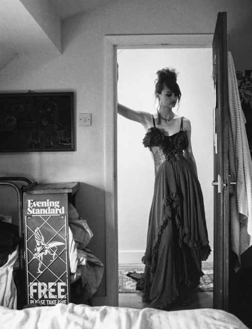 A young adult woman in a formal dress stands in a doorway inside a house, captured in black and white with a bed and newspaper stand nearby.