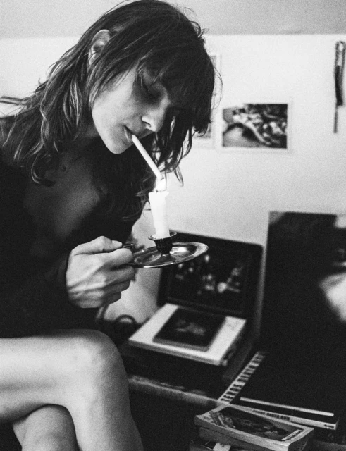 Monochrome portrait of a woman lighting a cigarette with a candle, sitting indoors with books and artwork in the background.