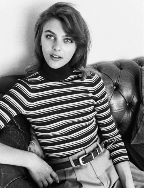 Portrait of a woman with shoulder-length wavy hair wearing a striped turtleneck and tan pants sitting on a leather couch.