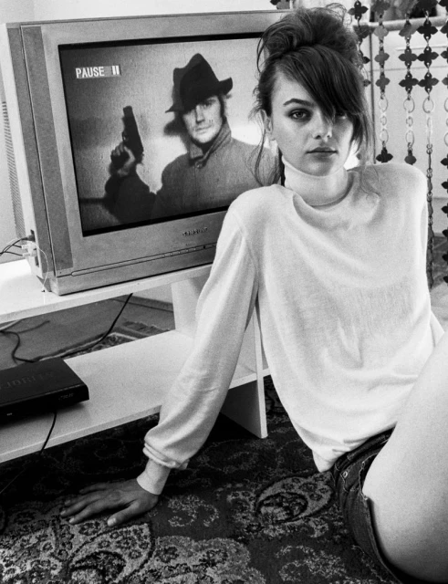 Black and white shot of a woman in a white sweater sitting in front of a retro television with an image of a man holding a gun.