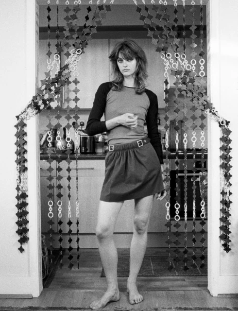 Full-length black and white shot of a woman standing in a doorway, wearing a t-shirt and skirt, with a beaded curtain backdrop.