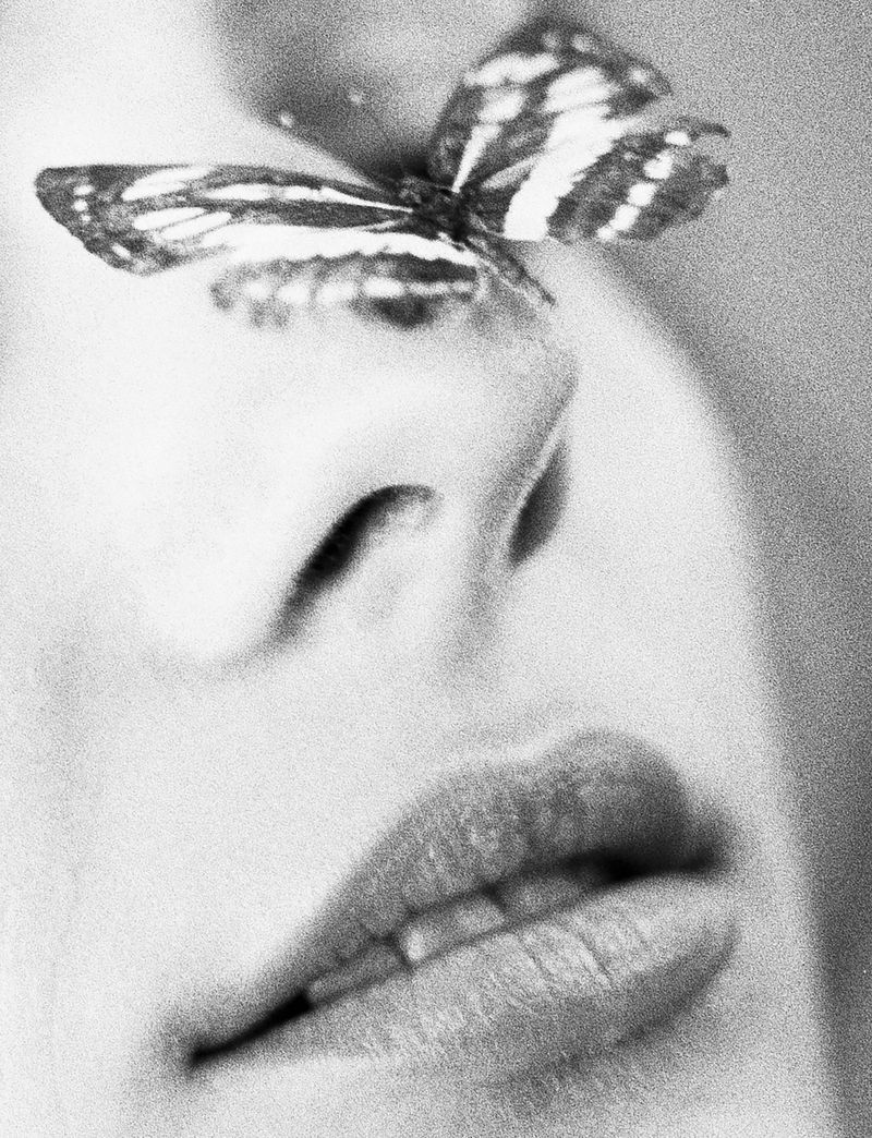 Close-up black and white shot of a woman's face with a butterfly perched on her nose, emphasizing beauty and nature.