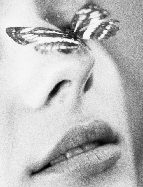 Close-up black and white shot of a woman's face with a butterfly perched on her nose, emphasizing beauty and nature.