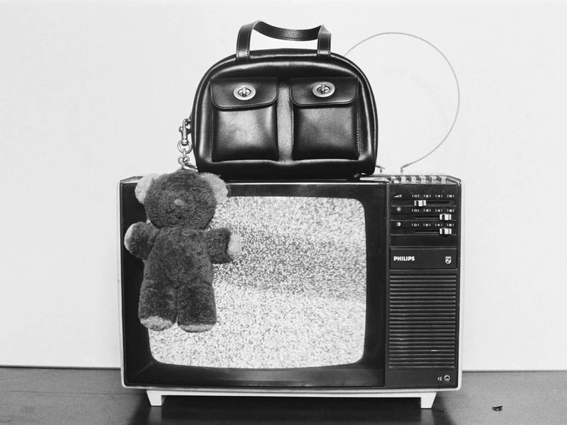 Black and white studio shot of a black handbag on an old television set with a plush teddy bear in front, against a white backdrop.