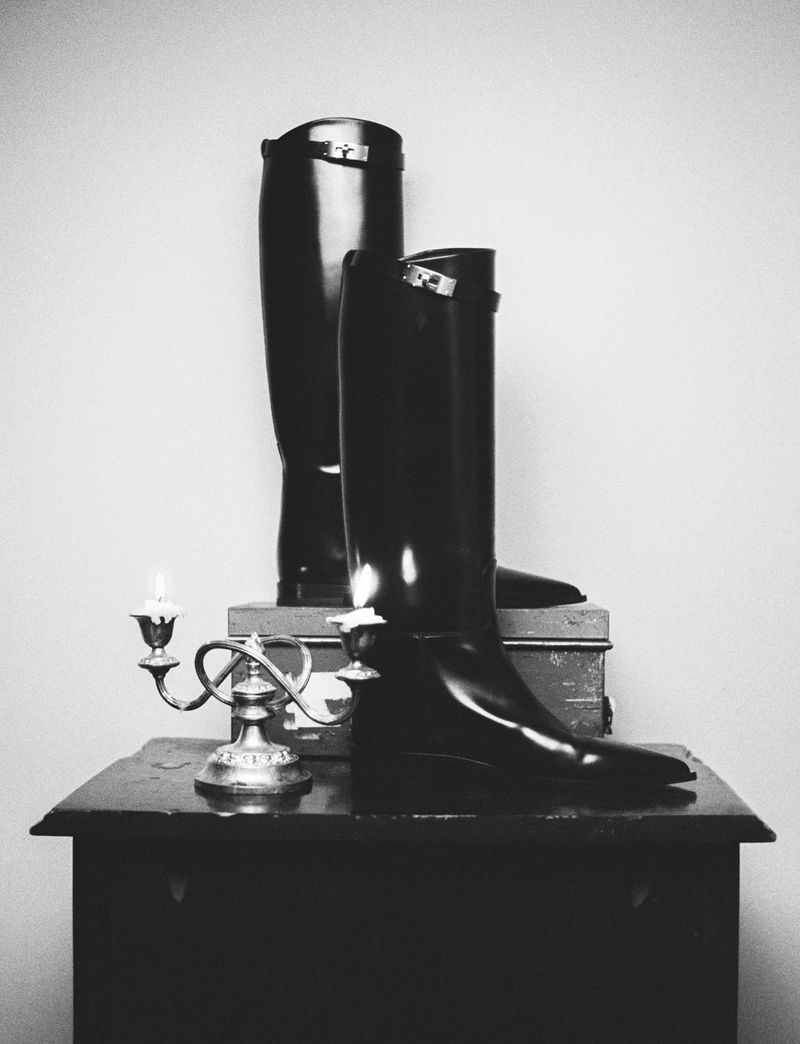 Monochrome still life featuring a pair of tall, shiny black riding boots displayed on a wooden table alongside a silver candelabra.