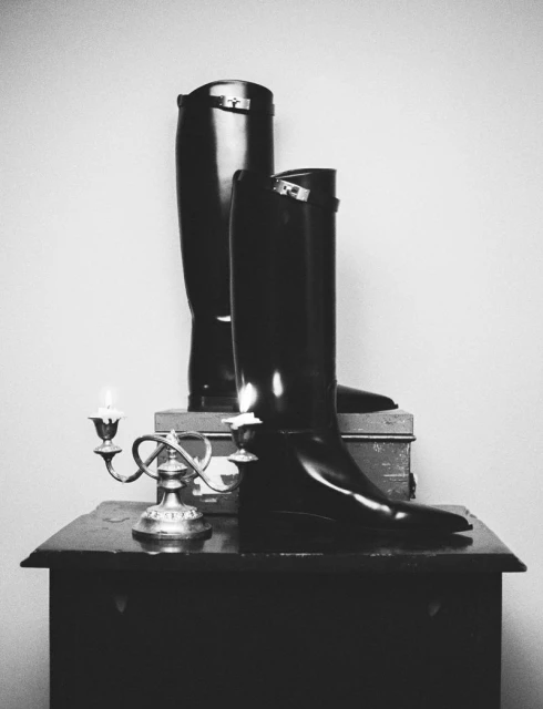 Monochrome still life featuring a pair of tall, shiny black riding boots displayed on a wooden table alongside a silver candelabra.