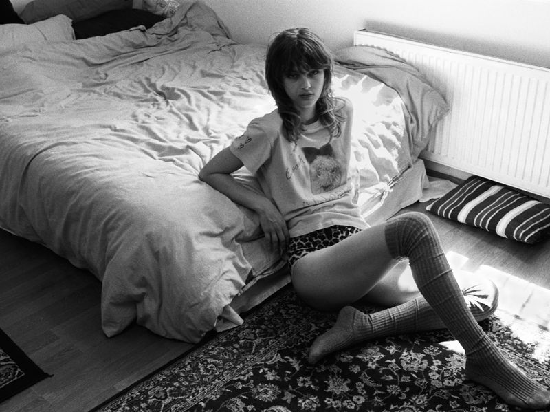 Monochrome portrait of a young adult female model sitting on the floor next to a bed, wearing knee socks, in an indoor setting.