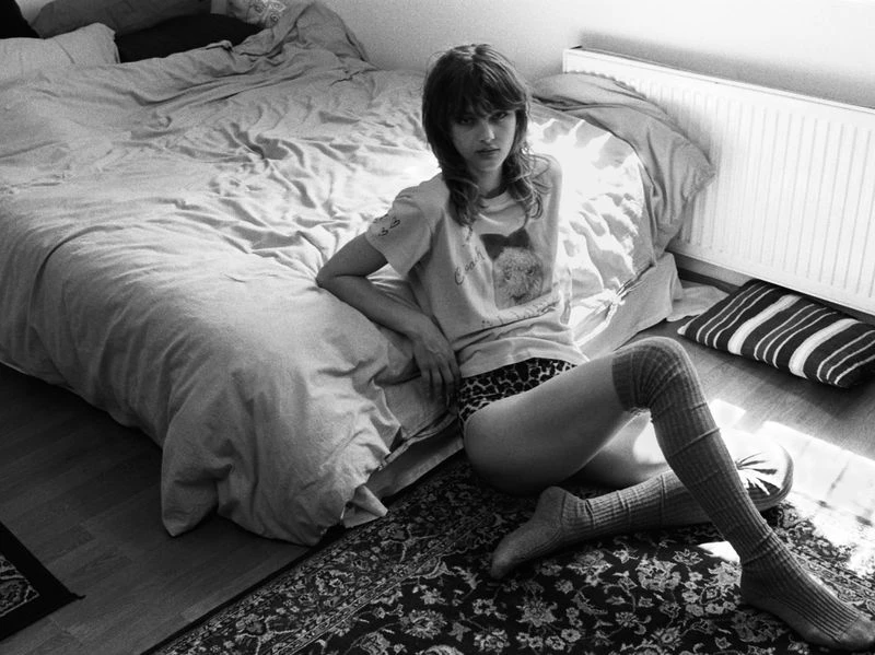 Monochrome portrait of a young adult female model sitting on the floor next to a bed, wearing knee socks, in an indoor setting.