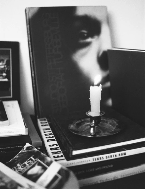 Monochrome photo of a lit candle on a silver plate, resting atop a stack of books, with a large book cover in the background.