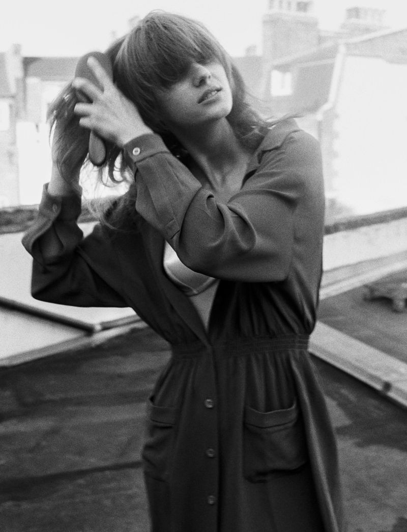 Black and white portrait of a woman on a rooftop brushing her hair, wearing a dress with a button-up front and pockets.