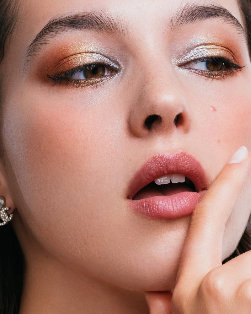 Close-up of a young woman with dramatic bronze and silver eye makeup, featuring a finger to the lips in a shushing gesture.