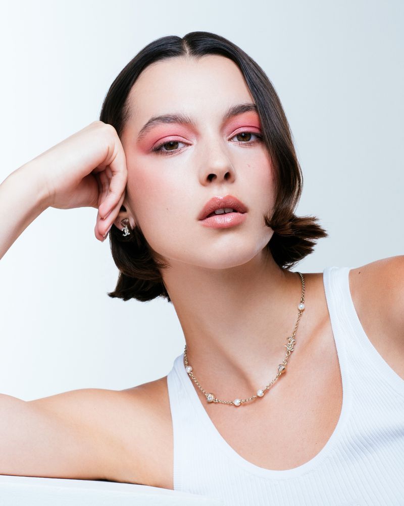 Close-up portrait of a woman with pink eyeshadow and short dark hair wearing a white tank top and pearl chain necklace.