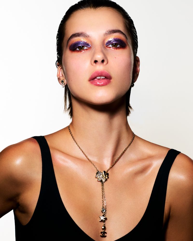 Portrait of a young adult woman with dramatic purple eyeshadow, slicked-back hair, and a Chanel necklace against a white backdrop.