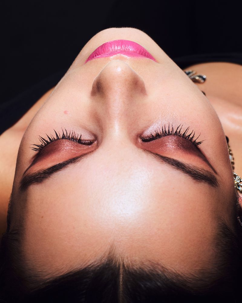 Close-up shot of a fair-skinned woman with closed eyes, defined eyebrows, pink lipstick, eye shadow, and earrings against a dark background.