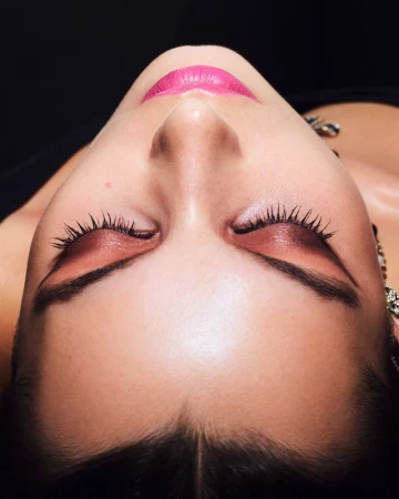 Close-up shot of a fair-skinned woman with closed eyes, defined eyebrows, pink lipstick, eye shadow, and earrings against a dark background.