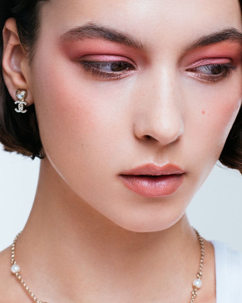 Close-up of a young woman with neutral makeup and rose eyeshadow, wearing gold earrings and a gold chain necklace with pearl beads.