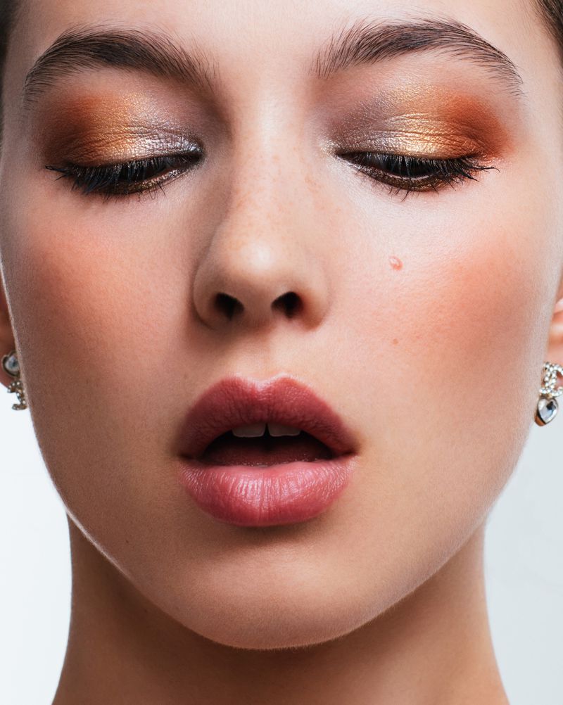 Close-up portrait of a young woman with closed eyes and bronze and gold eyeshadow, pink lips, freckles, and silver earrings.