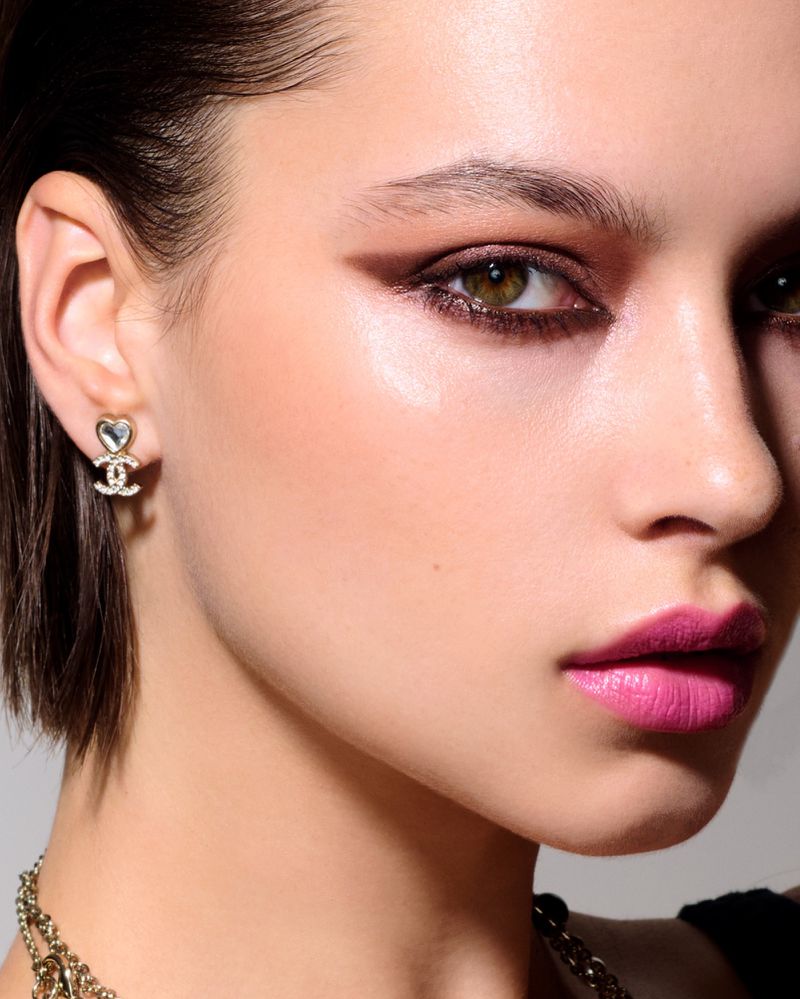 Close-up shot of a young woman with bold pink lipstick and dramatic eye makeup, featuring heart-shaped Chanel earrings in a beauty context.