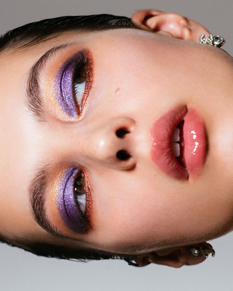 Close-up portrait of an adult female with vibrant eye makeup, including purple eyeshadow and glossy lips, against a plain background.