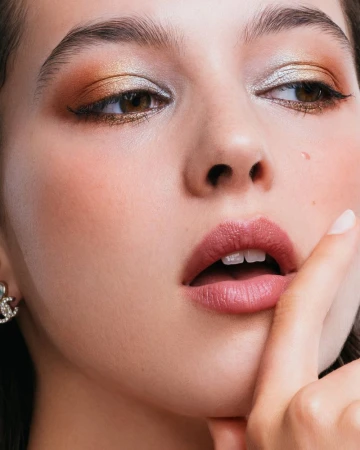 Extreme close-up beauty portrait of a woman with shimmering gold eyeshadow, pink lips, and a silver logo earring.
