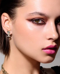 Close-up beauty portrait of a woman wearing pink lipstick, dark eye makeup, and CC logo heart earrings for Chanel Beauty.