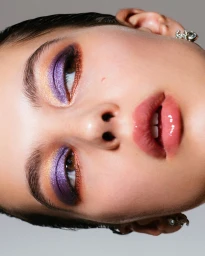 Close-up beauty portrait of a model with vibrant purple and gold eyeshadow and glossy pink lips against a neutral background.