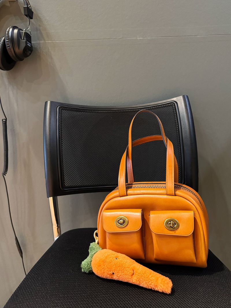 Orange leather handbag with pockets and a carrot keychain displayed on a black chair against a grey wall with headphones.