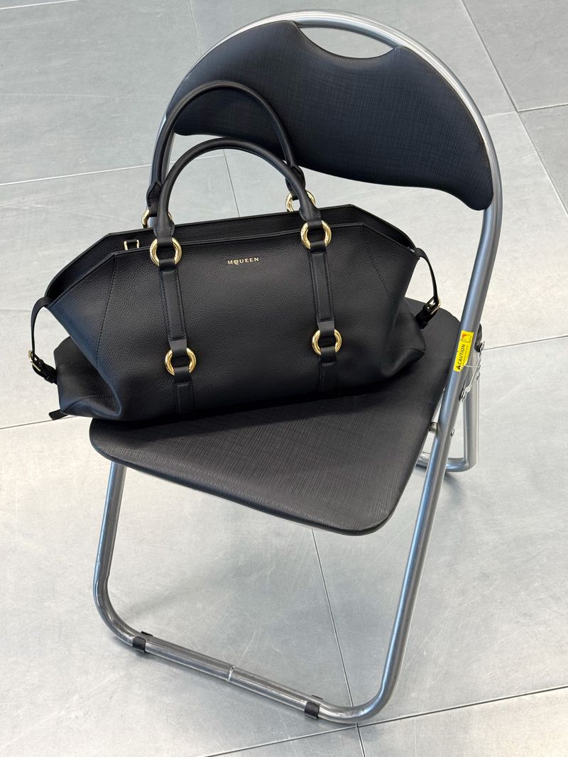 Black leather designer handbag with gold circular hardware resting on a black folding metal chair over grey tiled flooring.