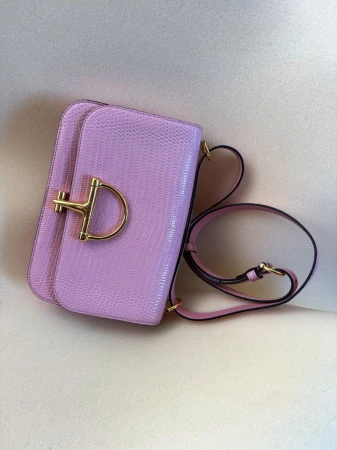 A light purple shoulder bag with a gold-toned clasp and strap on a beige surface, showcasing fashion accessory details.