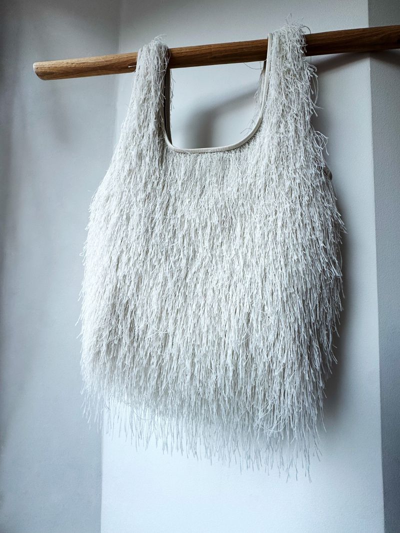 Fringed textile tote bag hanging from a wooden rod against a soft white wall, featuring a unique textured design and neutral tones.