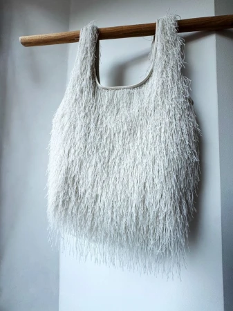 Fringed textile tote bag hanging from a wooden rod against a soft white wall, featuring a unique textured design and neutral tones.