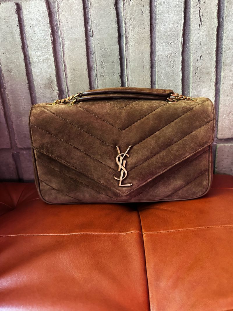 Brown suede YSL handbag with gold chain strap and logo on a orange leather surface in front of a gray textured background.