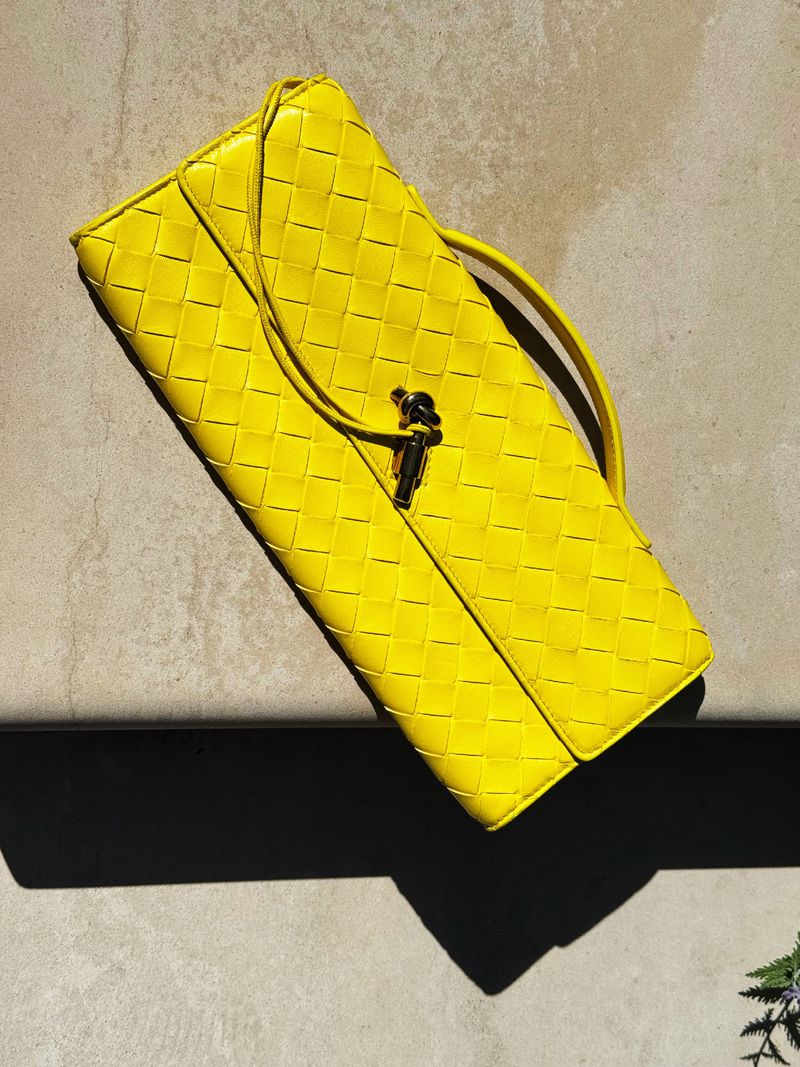 Bright yellow Bottega Veneta clutch bag with woven leather texture and metal clasp resting on a textured neutral surface in studio lighting.