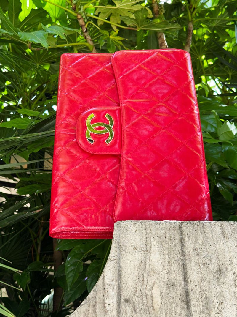 Bright red, quilted Chanel wallet on a weathered concrete surface against a backdrop of vibrant green foliage and natural light.