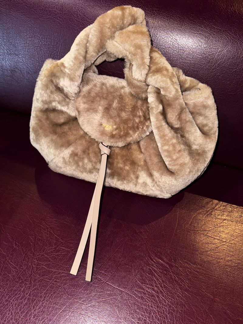 Tan faux fur purse with gathered top and thin straps on a burgundy background. Faux fur texture and neutral color palette.